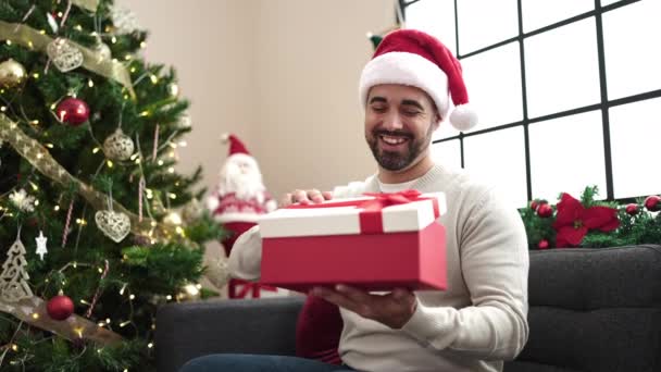 Jonge Spaanse man uitpakken geschenk met ongelukkige uitdrukking zitten op de bank door kerstboom thuis - Video