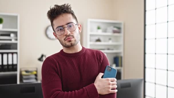 Jovem hispânico homem empresário de pé com braços cruzados gesto segurando smartphone no escritório - Filmagem, Vídeo