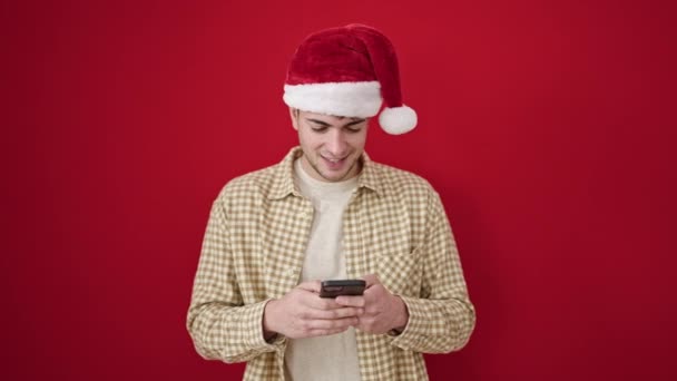 Νεαρός Ισπανόφωνος φοράει χριστουγεννιάτικο καπέλο χρησιμοποιώντας smartphone γιορτάζοντας πάνω από απομονωμένο κόκκινο φόντο - Πλάνα, βίντεο