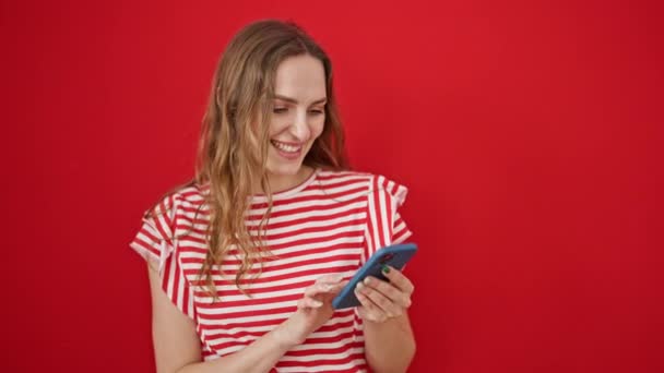 Mujer rubia joven usando teléfono inteligente sonriendo sobre fondo rojo aislado - Imágenes, Vídeo