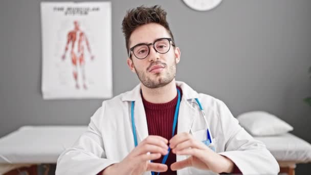 Joven médico hispano sonriendo haciendo gesto de corazón con las manos en la clínica - Metraje, vídeo