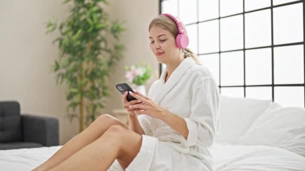 Mujer rubia joven escuchando música sentada en la cama bailando en el dormitorio - Imágenes, Vídeo