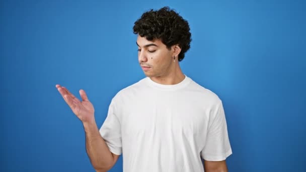 Jonge latijnse man glimlachend wijzend naar de zijkant presenteren over geïsoleerde blauwe achtergrond - Video