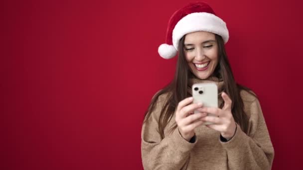 Gyönyörű spanyol nő visel karácsonyi kalap segítségével okostelefon felett elszigetelt piros háttér - Felvétel, videó