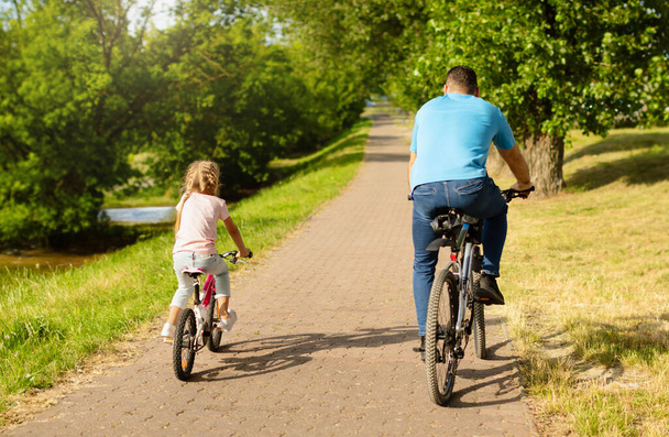 Achteraanzicht van vader van middelbare leeftijd en zijn dochtertje in casual kleding fietsen rijden in het park, wandelen en tijd samen doorbrengen in het weekend, genieten van goed zonnig weer. Lichamelijke activiteit, recreatie - Foto, afbeelding