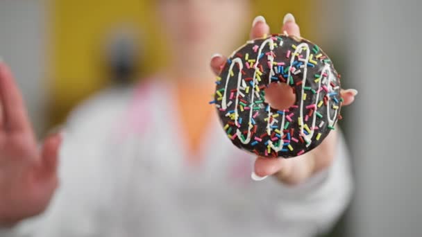 Joven mujer rubia dietista sosteniendo donut haciendo gesto de parada con la mano en la clínica - Metraje, vídeo