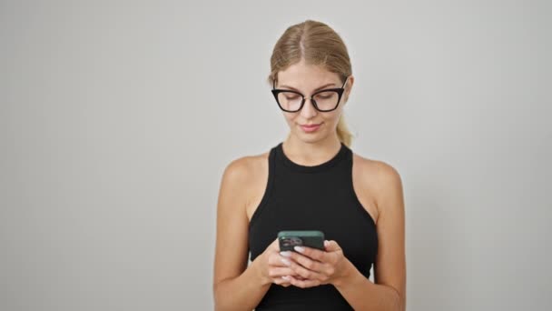 Молодая блондинка с помощью смартфона смотрит в сторону улыбаясь на изолированном белом фоне - Кадры, видео