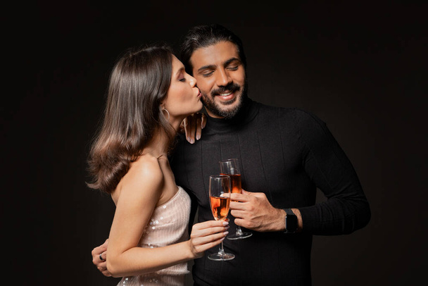 シャンパンのガラスを保持するエレガントな衣装で幸せな美しい若い女性,抱擁し,彼女の男性にキス,カップルを祝うXmas,新年,記念日,黒い背景での誕生日,コピースペース - 写真・画像