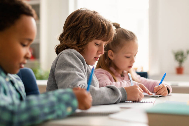Διαφορετική γραφή μαθητή κατά τη διάρκεια του μαθήματος στο γραφείο στην τάξη στο δημοτικό σχολείο, κάθεται στη σειρά, κάνει εργασίες δοκιμής. Παιδιά που γράφουν σημειώματα στην τάξη - Φωτογραφία, εικόνα