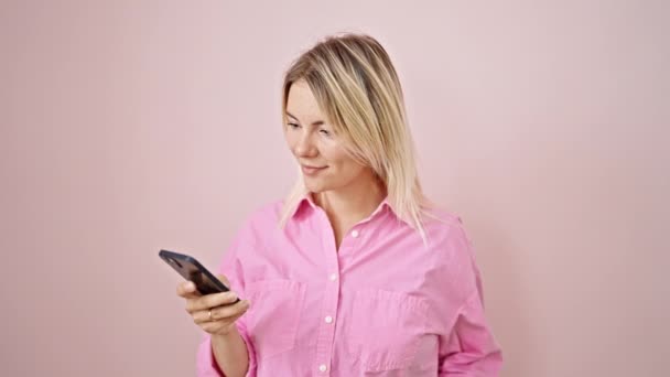 Mladá blondýna žena pomocí smartphone usmívá přes izolované růžové pozadí - Záběry, video