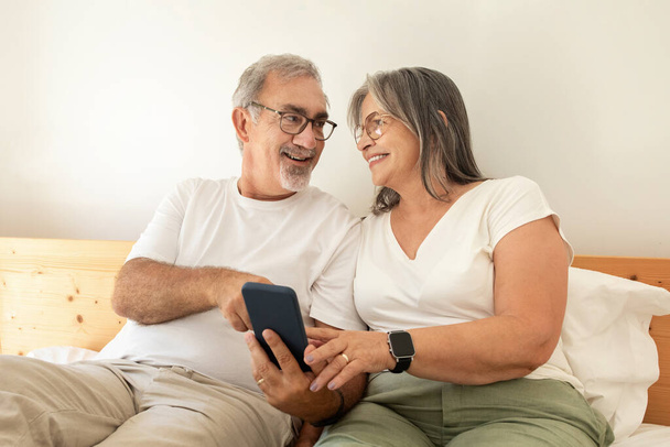 Ευτυχισμένοι ηλικιωμένοι Ευρωπαίοι σύζυγοι βλέπουν βίντεο στο τηλέφωνο, κουβεντιάζουν, κάθονται στο κρεβάτι στο εσωτερικό της κρεβατοκάμαρας. Ξεκούραση και χαλάρωση μαζί, photo app και συσκευή, διασκέδαση στον ελεύθερο χρόνο στο σπίτι - Φωτογραφία, εικόνα