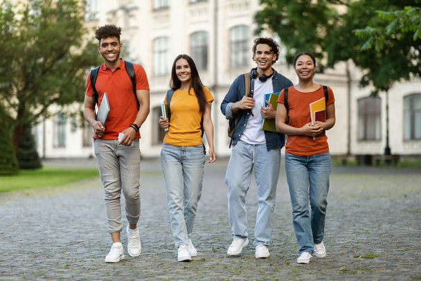 Retrato de felices amigos alegres de la universidad caminando al aire libre después de clases, jóvenes estudiantes multiétnicos positivos llevando libros de trabajo y mochilas, sonriendo y mirando la cámara mientras van al campus - Foto, Imagen