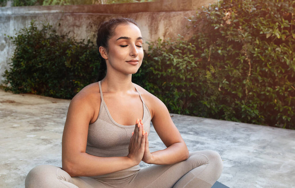 Спокойная молодая арабская спортсменка в спортивной одежде с закрытыми глазами практикует йогу, медитирует в позе лотоса, наслаждается дыхательными упражнениями, на свежем воздухе в летнее утро. Спорт, фитнес, только тренировка - Фото, изображение
