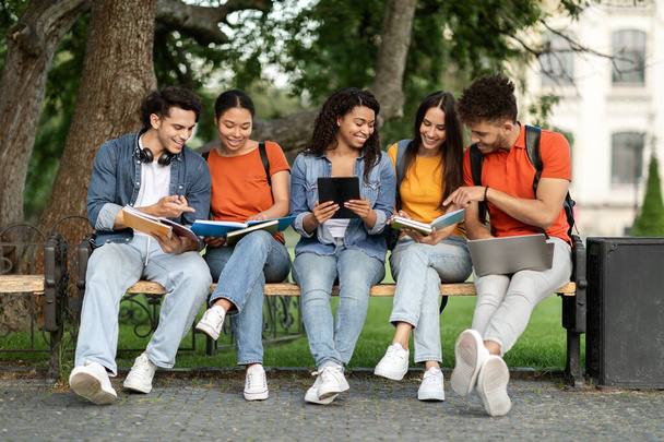 Ομάδα πολυεθνικών φοιτητών σπουδάζουν μαζί σε εξωτερικούς χώρους στην πανεπιστημιούπολη, ευτυχισμένοι νέοι φίλοι από το κολέγιο που κάθονται στον πάγκο έξω, προετοιμάζονται για τις εξετάσεις, χρησιμοποιώντας ψηφιακά tablet, laptop και βιβλία εργασίας - Φωτογραφία, εικόνα