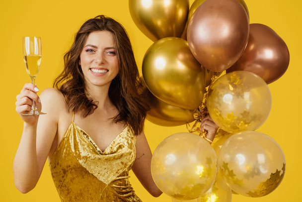 Ευτυχισμένη όμορφη κυρία με κομψό φόρεμα κρατώντας αερόστατα αέρα και ένα ποτήρι σαμπάνια, χαμογελώντας στην κάμερα σε κίτρινο φόντο. Ενθουσιασμένη γυναίκα σε πάρτι γενεθλίων - Φωτογραφία, εικόνα