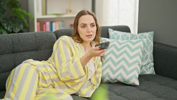 Young blonde woman watching tv lying on sofa at home - Filmmaterial, Video
