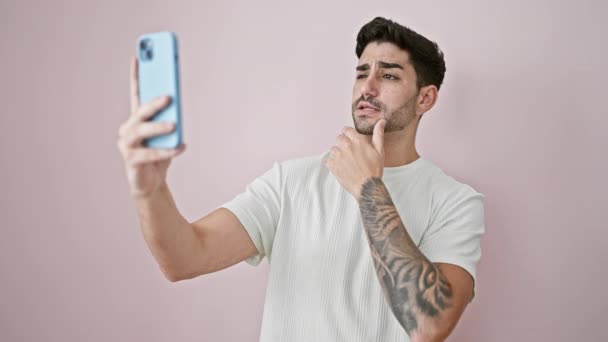 Giovane uomo ispanico sorridente fiducioso facendo selfie dallo smartphone su sfondo rosa isolato - Filmati, video
