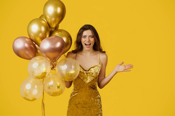 Tyytyväinen tunteellinen nainen, jolla on ilmapalloja, katselee kameraa jännityksellä, seisoo eristettynä keltaisella studiotaustalla. Syntymäpäiväjuhlat ja loma juhla - Valokuva, kuva