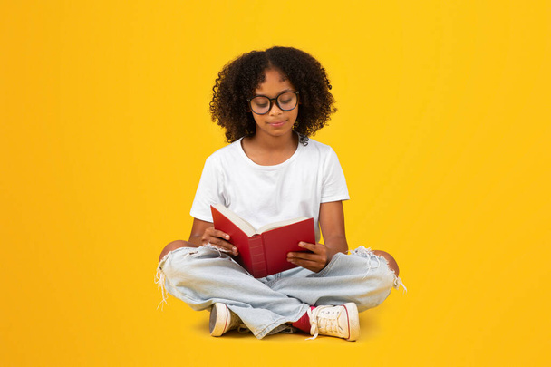 Adolescente intelligente sérieuse afro-américaine en t-shirt blanc, livre de lecture de lunettes, assise sur le sol, isolée sur fond orange, studio. Travail à domicile et repos, passe-temps, études, connaissances, éducation scolaire - Photo, image