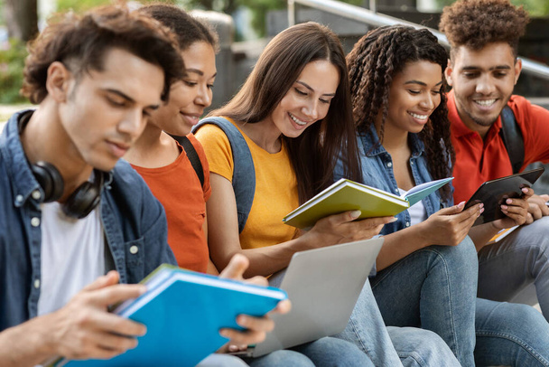 若い多民族のグループ 屋外で一緒に勉強,5人の学生が本を読み,外で座っている間にデジタルタブレットを使用して,試験のために準備するか,クラスを閉じる - 写真・画像