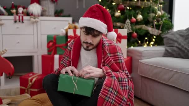 Joven hombre hispano desempacando regalo sentado junto al árbol de Navidad en casa - Imágenes, Vídeo