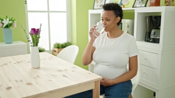 Молода вагітна жінка п'є склянку води, сидячи на столі в їдальні
 - Кадри, відео