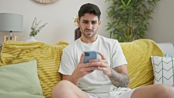 Молодий іспаномовний чоловік використовує смартфон, сидячи на дивані, дивлячись засмучений вдома - Кадри, відео
