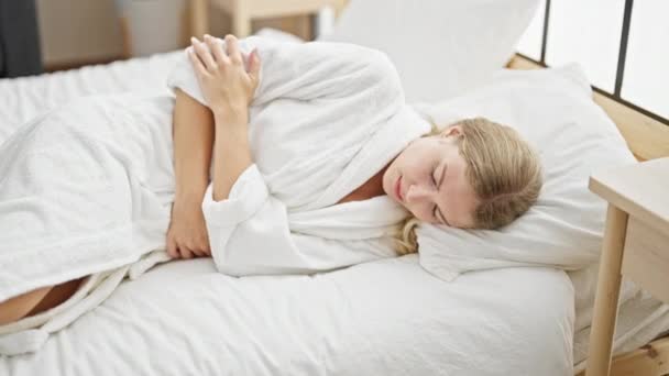 Νεαρή ξανθιά γυναίκα που υποφέρει από εμμηνορροϊκό πόνο ξαπλωμένη στο κρεβάτι στο υπνοδωμάτιο - Πλάνα, βίντεο