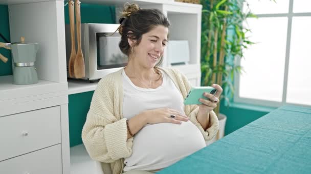 Mujer embarazada joven viendo vídeo en el teléfono inteligente tocando el vientre en el comedor - Metraje, vídeo