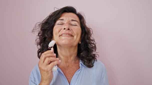 Střední věk hispánská žena usmívající se sebevědomá masáž obličeje s kožní váleček přes izolované růžové pozadí - Záběry, video