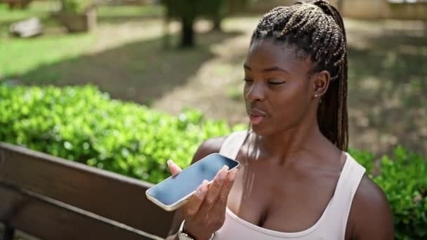 Αφροαμερικανή γυναίκα στέλνει φωνητικό μήνυμα με smartphone κάθεται στον πάγκο στο πάρκο - Πλάνα, βίντεο