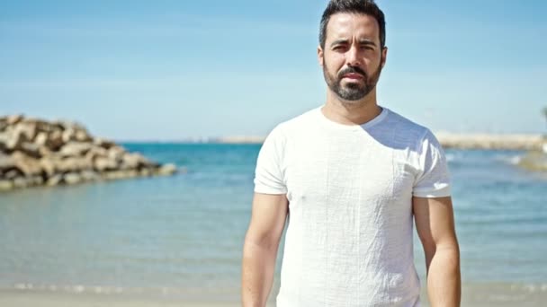 Молодий іспаномовний чоловік дивиться в бік з серйозним виразом на пляжі - Кадри, відео