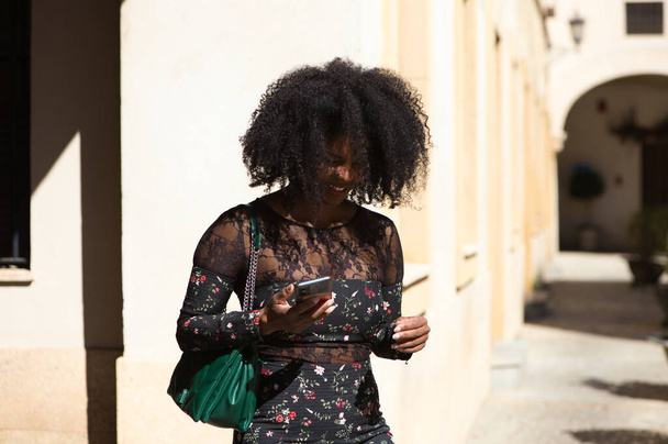 Portret van een zwarte vrouw, jong en mooi met afro haar en zwarte jurk met bloemen en groene handtas wandelingen en glimlach, de vrouw houdt mobiele telefoon in haar hand - Foto, afbeelding