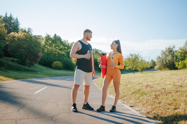 Two young adults, both athletes, a man, and a woman, in sportswear, engaging in conversation and enjoying the outdoors after their workout. while the woman is holding a fitness mat. - Фото, изображение