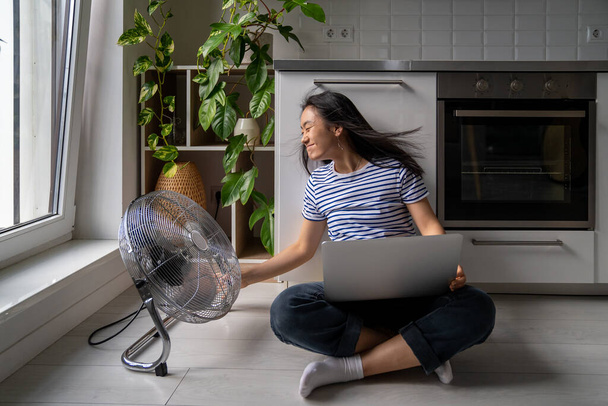 Femme transformée ventilateur électrique puissant assis jambes croisées sur le sol dans la cuisine moderne travaillant sur ordinateur portable refroidissement chaleur d'été. Femme asiatique freelance combats effets réchauffement climatique avec ventilateur de sol. - Photo, image
