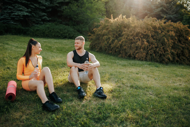 Two athletes, a man and woman, wearing sportswear, enjoying each others company outside after their workout. The man holds a water bottle, and the woman is holding a fitness mat. - Foto, Bild