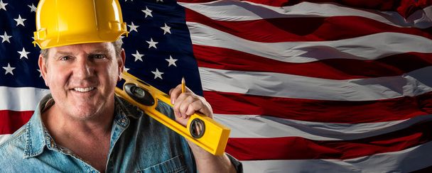 Contratista masculino que usa el Hardhat amarillo en blanco sobre el banner de fondo de bandera estadounidense ondeante. - Foto, imagen