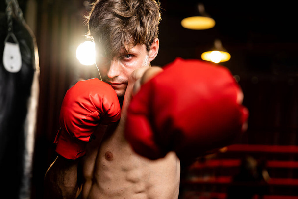 Boxkämpfer ohne Hemd posiert, kaukasischer Boxer schlägt seine Faust vor der Kamera in aggressiver Haltung und bereit, im Fitness-Studio zu kämpfen, im Hintergrund Trettasche und Boxausrüstung. Impulse - Foto, Bild