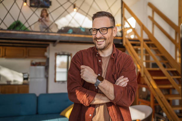 Egy felnőtt fehér férfi szakállal és szemüveggel otthon áll, boldog mosollyal másolja a helyet. - Fotó, kép