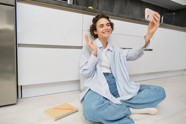 Portret van jonge vrouw videochats, neemt vlog op terwijl ze op de keukenvloer zit. Schattig meisje neemt selfie op smartphone. - Foto, afbeelding
