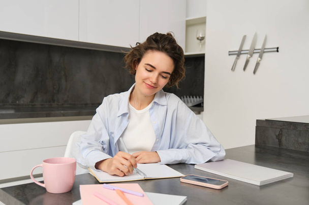 Retrato de una joven que trabaja desde casa, anotando información en el cuaderno, tomando notas, sentada en la cocina y estudiando, estudiante haciendo la tarea. - Foto, imagen