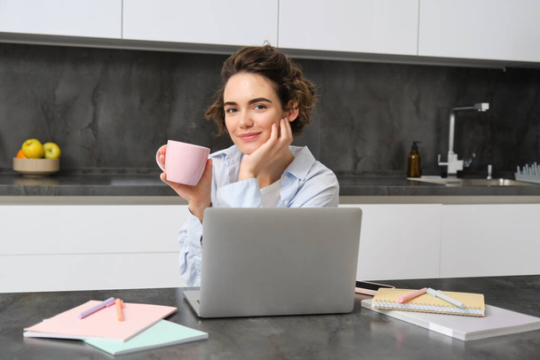 Samozatrudniona kobieta pracuje w domach, siedzi z laptopem i pije kawę w kuchni, otoczona dokumentami. Studentka uczy się zdalnie, projektuje na komputerze, trzyma kubek. - Zdjęcie, obraz