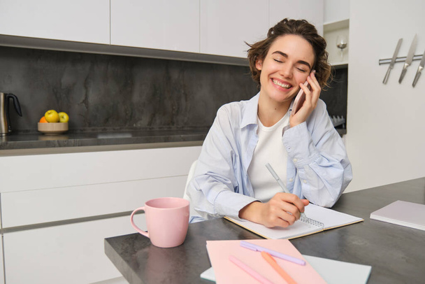 Portret szczerej, uśmiechniętej młodej kobiety, rozmawiającej przez telefon komórkowy, śmiejącej się podczas rozmowy na smartfonie, spisującej, notującej w notatniku, siedzącej w kuchni w domu. - Zdjęcie, obraz