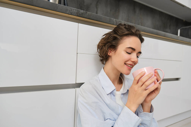 Koncept krásy a žen. Uvolněná mladá žena si vychutnává šálek čaje. Dívka sedí na podlaze v kuchyni s růžovým hrnkem, pije kávu a usmívá se radostí. - Fotografie, Obrázek
