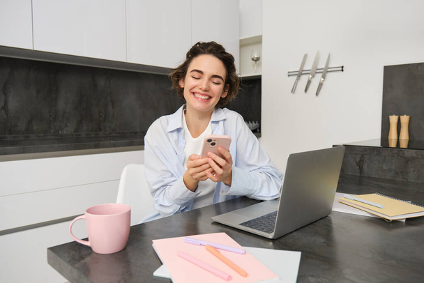 Зображення молодої жінки, яка використовує свій смартфон вдома. Дівчина сидить з мобільним телефоном на кухні і посміхається. - Фото, зображення