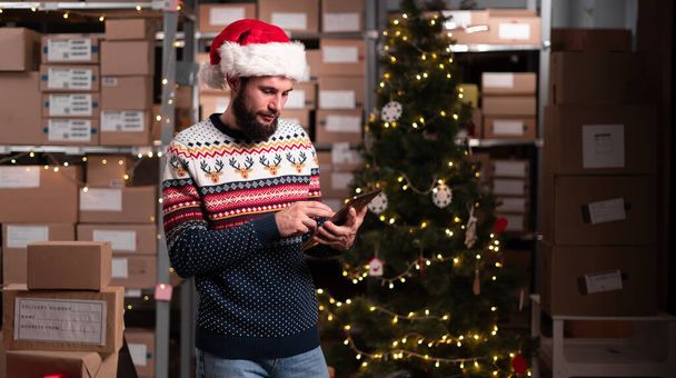 Miespuolinen varastotyöntekijä, joka työskentelee jouluna digitaalisen tabletin avulla varastossa. Joulun toimitus käsite - Valokuva, kuva
