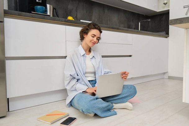 Freelance and lifestyle concept. Young woman sits on floor with laptop, works from home, sets up her remote workspace in kitchen, does homework. - Photo, Image