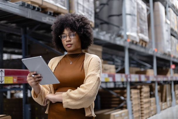 Jeune afro-américaine femelle regardant la caméra travaillant dans un entrepôt en utilisant l'inspection numérique de contrôle de tablette sur les étagères.. - Photo, image