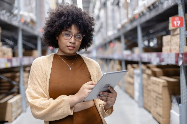 Young African American female looking at camera working in warehouse using digital tablet checking inspection on shelves.woman worker check stock inspecting in storage logistic factory. - Photo, Image