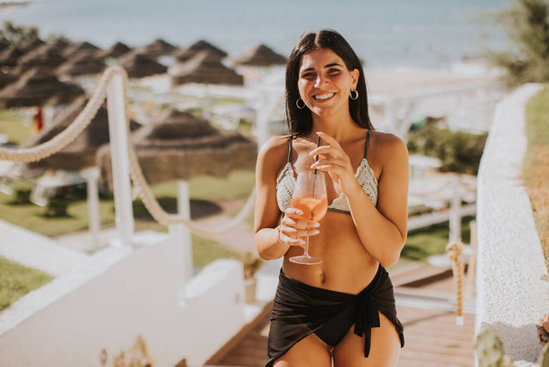 Посміхаючись молода жінка в Бікіні насолоджуючись відпусткою на пляжі під час пиття коктейлю - Фото, зображення